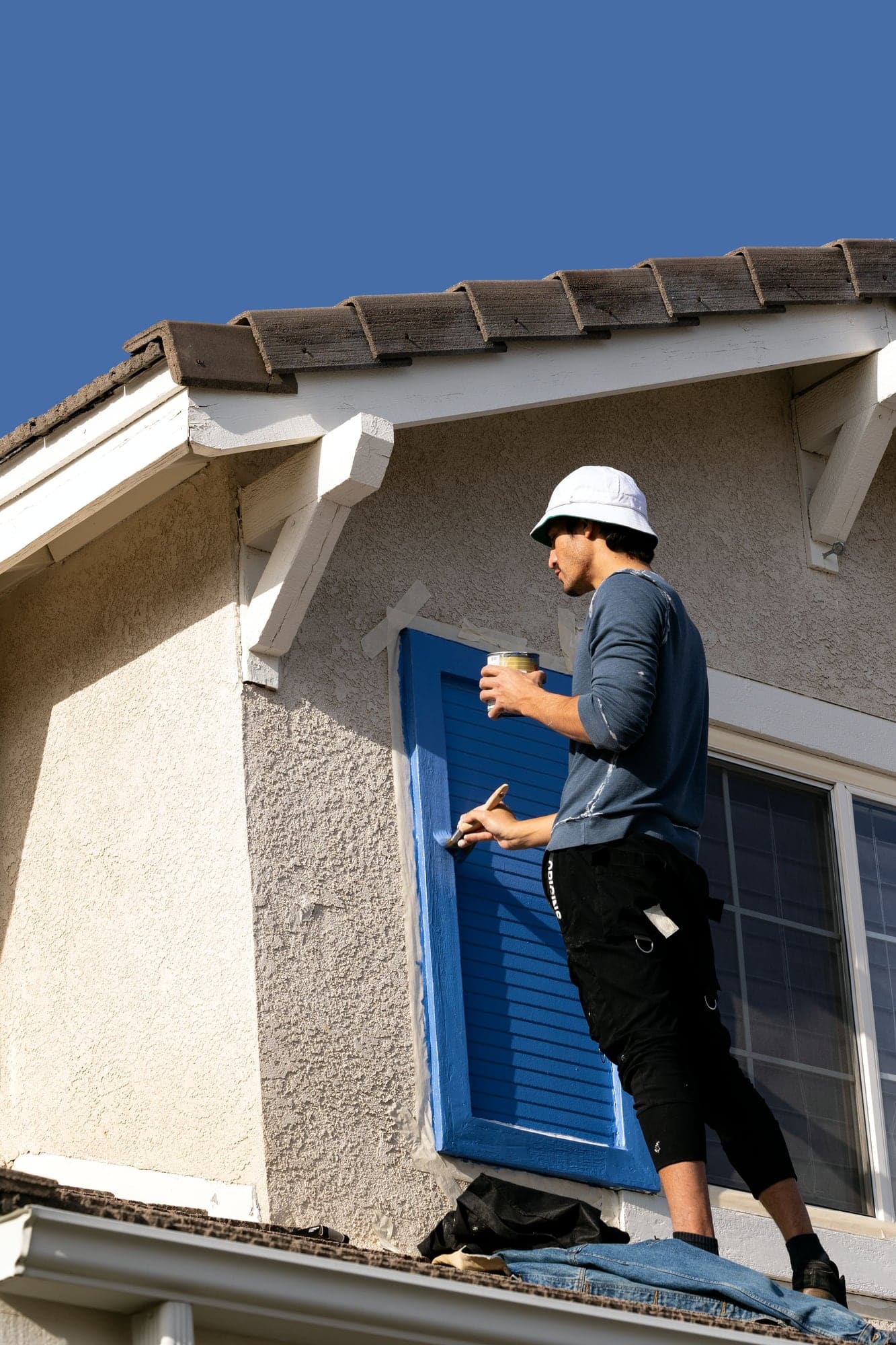 Peintre sur un échafaudage repeignant des volets bleus sur la façade crépie d’une maison.